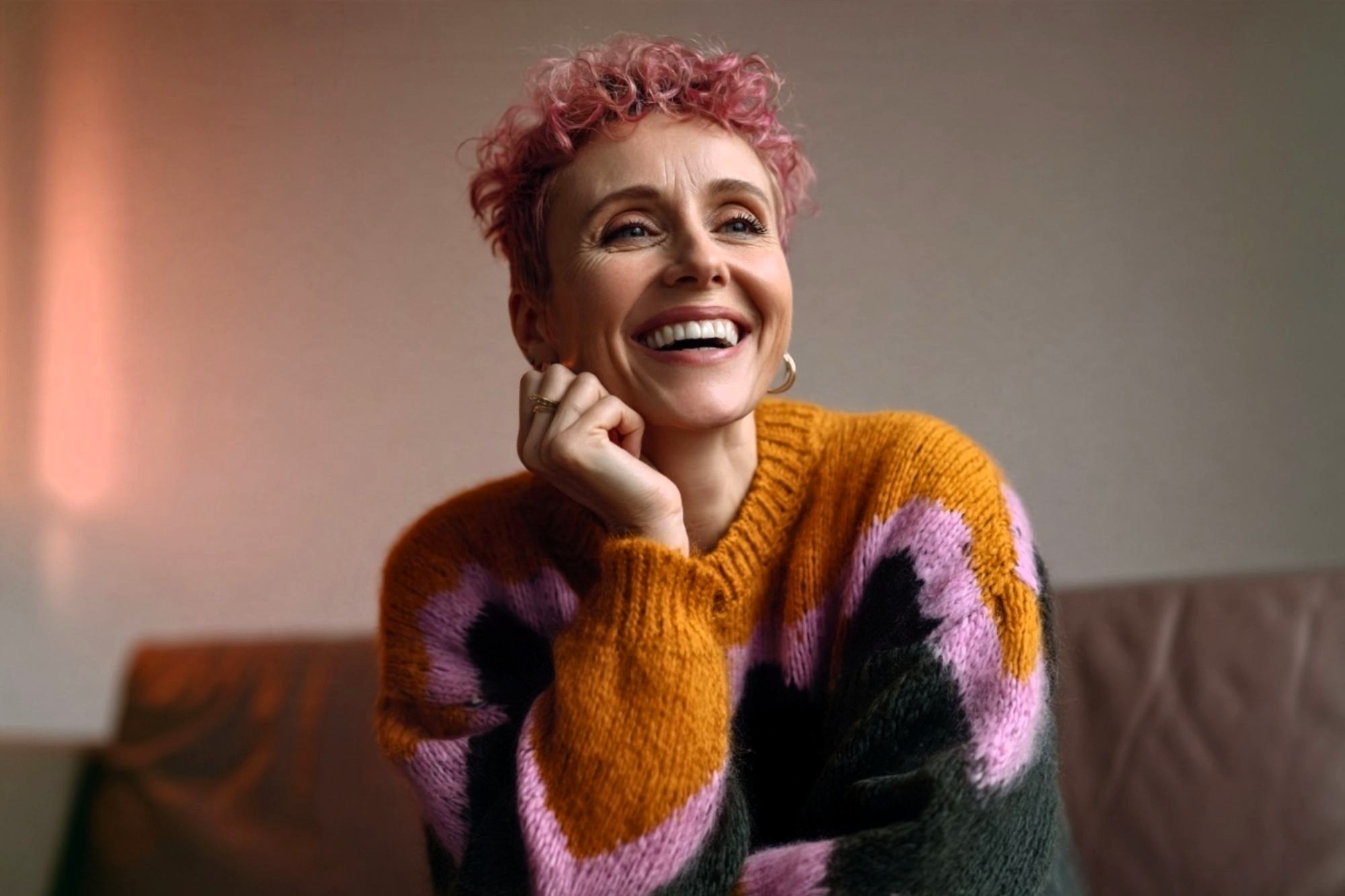 A smiling person with short, pink curly hair wears a chunky knit sweater with large blocks of orange, pink, and dark grey.