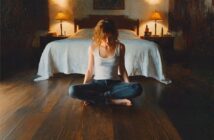 A woman sitting cross-legged on a wooden floor in front of a bed with two bedside lamps.