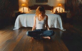 A woman sitting cross-legged on a wooden floor in front of a bed with two bedside lamps.