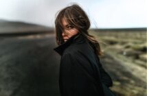 A person with long hair and a black coat stands on a deserted road, looking back over their shoulder. The wind blows their hair across their face, and the background is blurred and muted.