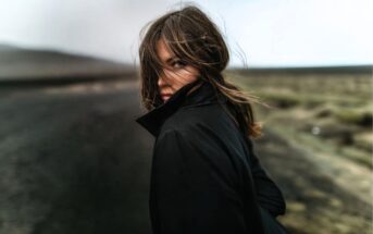 A person with long hair and a black coat stands on a deserted road, looking back over their shoulder. The wind blows their hair across their face, and the background is blurred and muted.