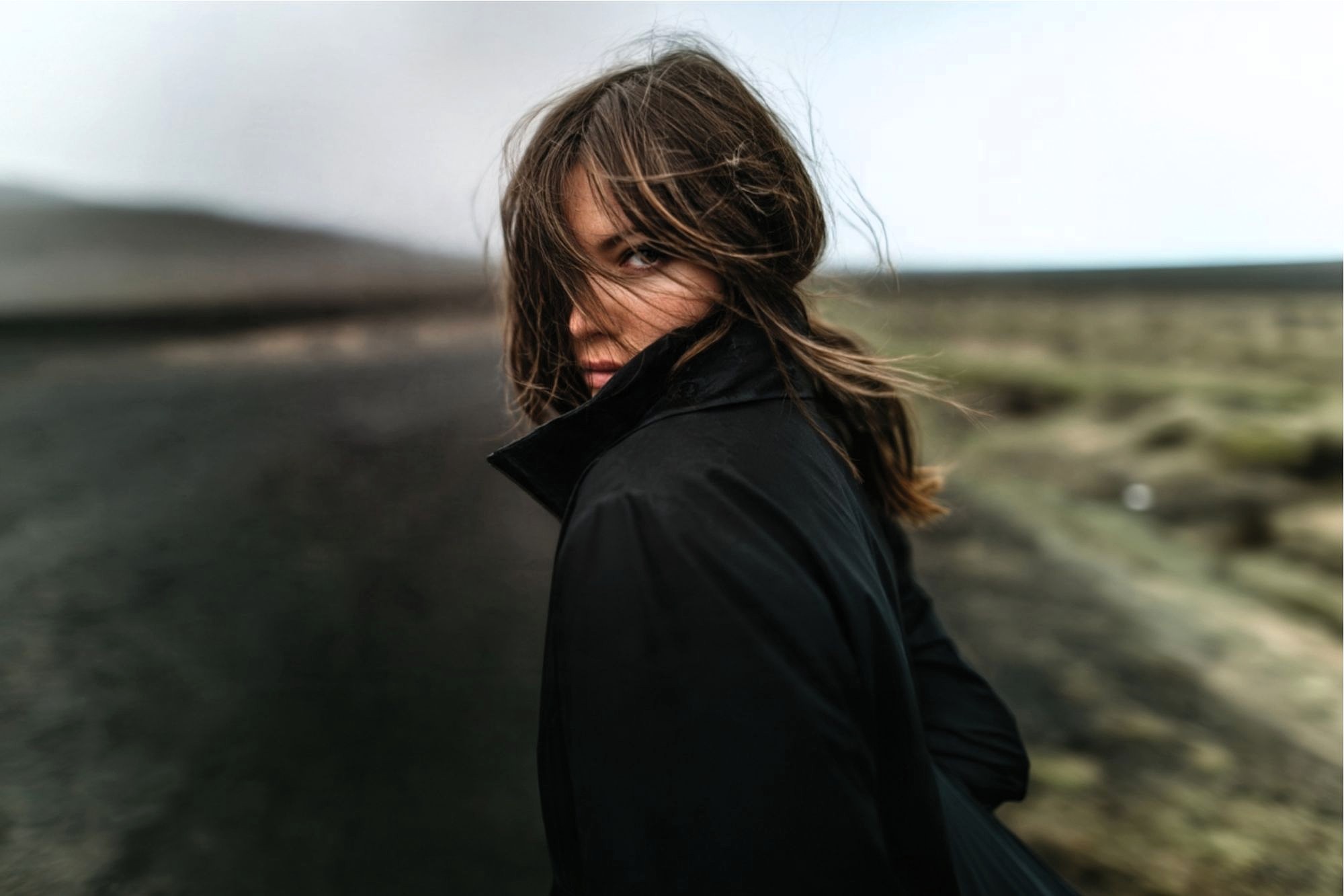 A person with long hair and a black coat stands on a deserted road, looking back over their shoulder. The wind blows their hair across their face, and the background is blurred and muted.