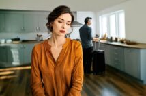 A woman in an orange shirt looks sad in a kitchen while a man with a suitcase stands by the window with his back turned, suggesting a tense or emotional moment.