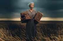 An older woman with gray hair stands in a windy field at dusk, wearing a patterned dress and holding three vintage suitcases, looking determined.