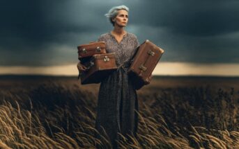 An older woman with gray hair stands in a windy field at dusk, wearing a patterned dress and holding three vintage suitcases, looking determined.