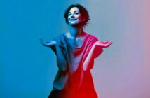 Smiling woman with dark hair, wearing a loose shirt, holds her hands up playfully. The image has a vibrant blue and pink gradient lighting effect.