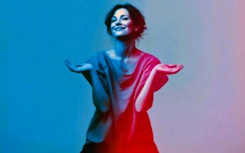 Smiling woman with dark hair, wearing a loose shirt, holds her hands up playfully. The image has a vibrant blue and pink gradient lighting effect.
