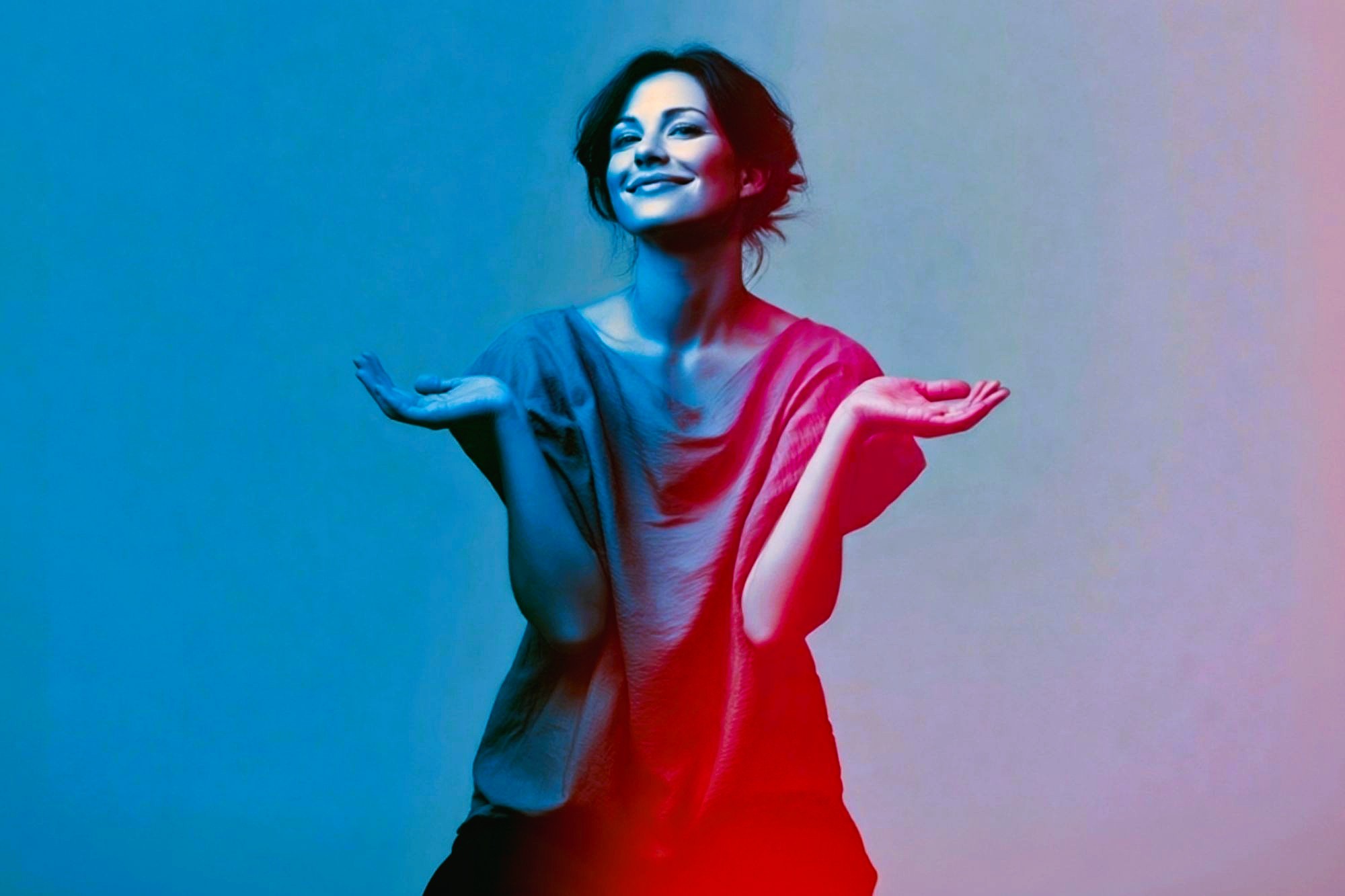 Smiling woman with dark hair, wearing a loose shirt, holds her hands up playfully. The image has a vibrant blue and pink gradient lighting effect.