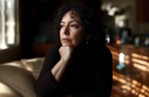 A woman with dark, curly hair sits with her hand on her chin, looking off to the side with a pensive expression.
