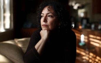 A woman with dark, curly hair sits with her hand on her chin, looking off to the side with a pensive expression.