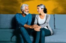 A middle-aged man and woman sitting on a sofa, looking at each other and holding hands.